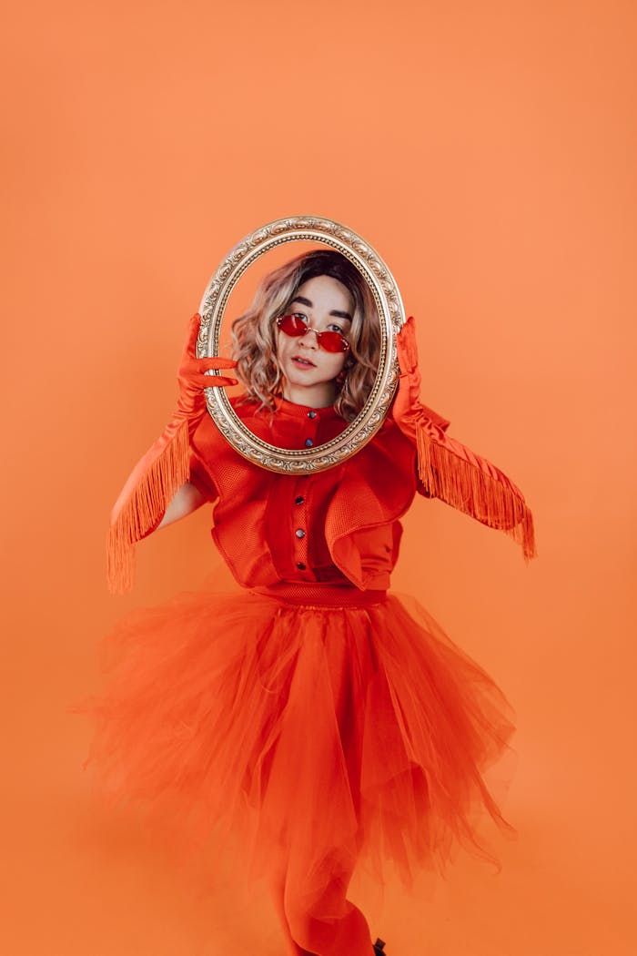 A person in an orange costume holding an ornate mirror against an orange background.